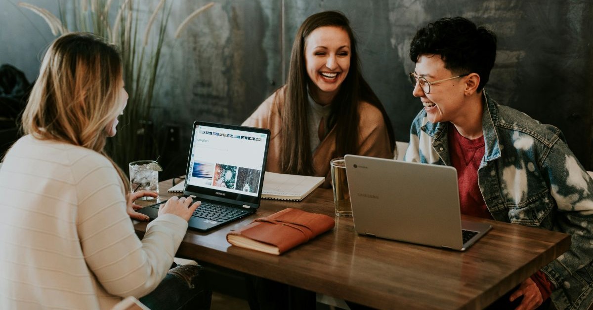 3 people sat at a table with their laptops smiling and laughing, having fun.