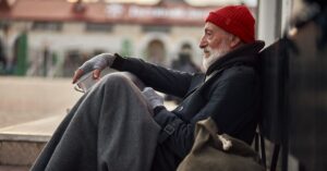 Side view of male on street begging, waiting help by citizens. Senior homeless man in old clothes sit on floor in cold weather.
