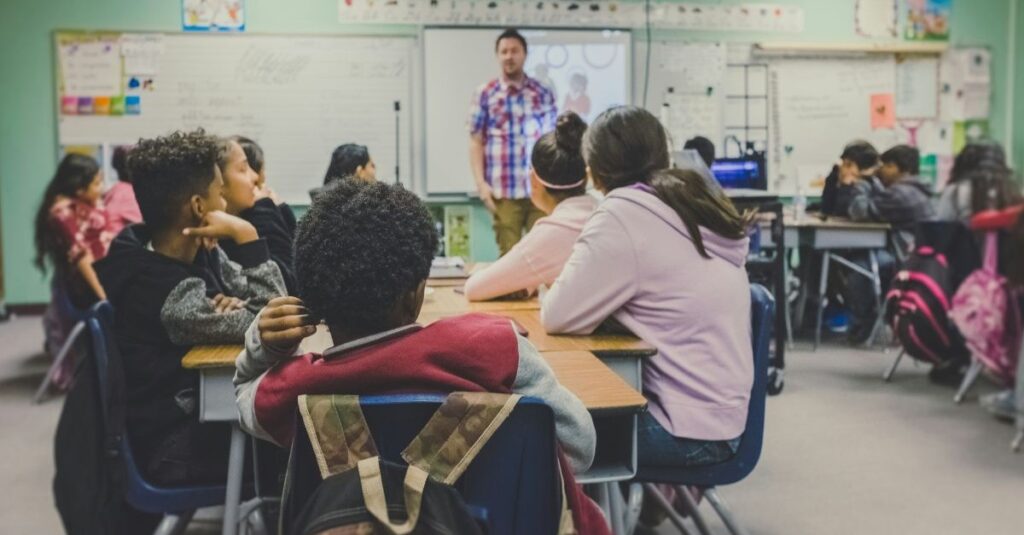 A classroom with a teacher at the front of the class talking to the children who are sat in their seats.