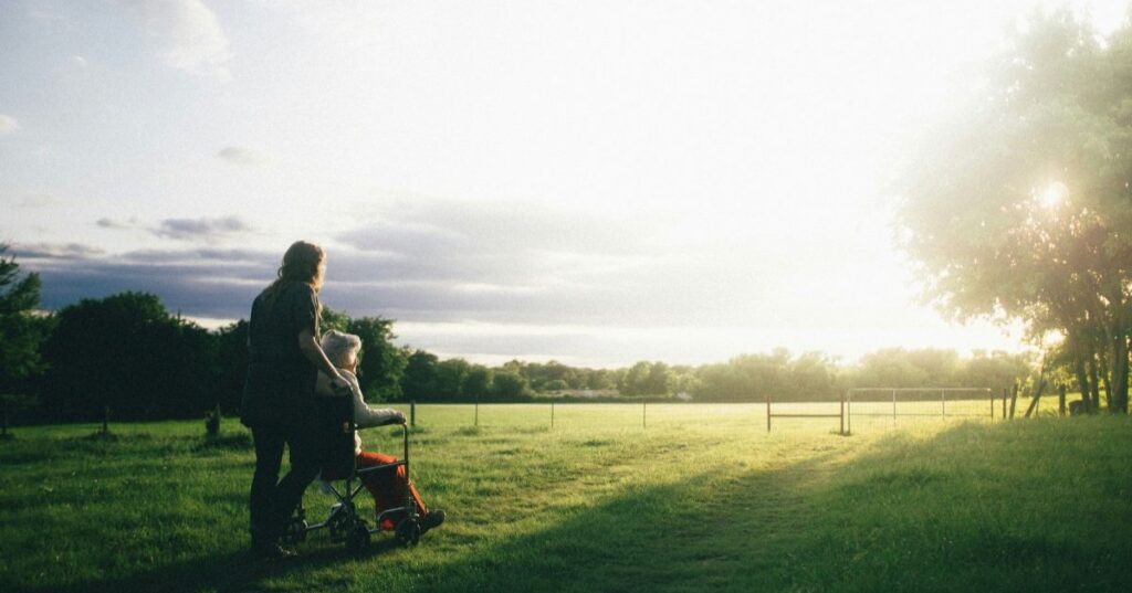 Woman walking, pushing an elderly woman in a wheelchair through the fields with the sun shining.