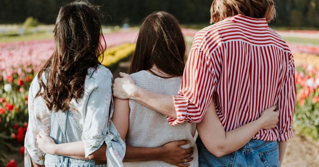 Three women stood in a flower field facing away from the camera, holding their arms around each other's backs, supporting each other.