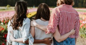 Three women stood in a flower field facing away from the camera, holding their arms around each other's backs, supporting each other.