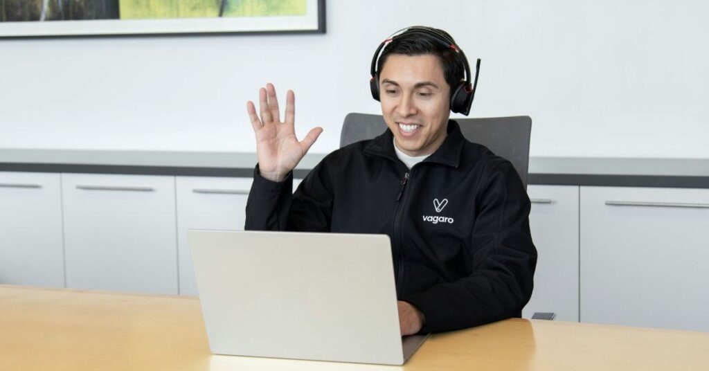 A man sat at his desk with his laptop on a call with a customer. He is holding his hand up, waving and smiling.